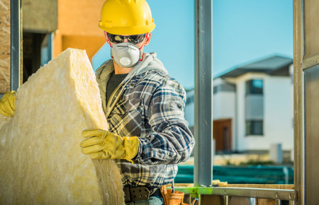 A man in a helmet and gloves lifts insulation material at a building site during the day.の写真素材