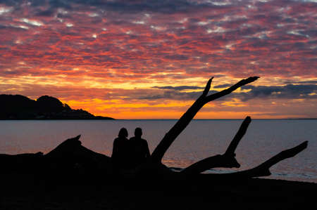 Couple sitting on a beached log enjoying each others company while watching the sunset.の写真素材