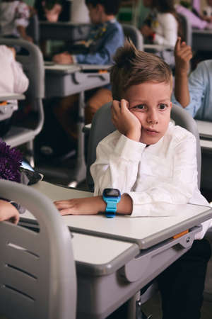 Pupil of primary school in uniform sitting in classroom. Upset and boring caucasian 6 years boy....