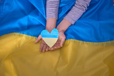 Child hands holding yellow-blue paper hearts on Ukrainian flag background. Kids support Ukraine,...