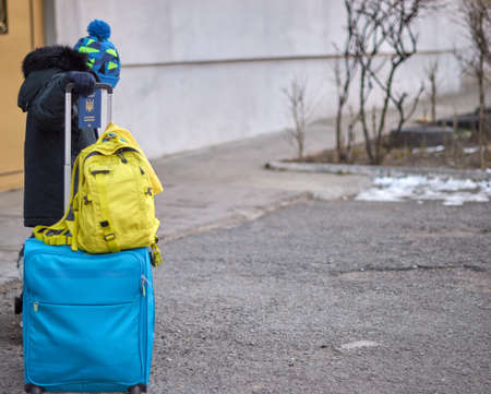 Evacuation of civilians, sad child with the flag of Ukraine. Refugee family from Ukraine crossing the border. Hand holding a passport above the luggage with yellow-blue flag. Stop war, support Ukraineの写真素材