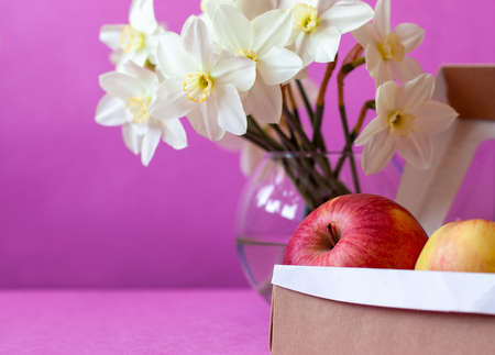 Red apple in a craft box and a bouquet of daffodils in a vase on pink backgroundの写真素材