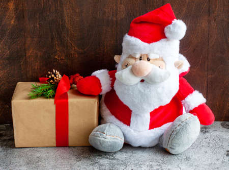 A toy Santa Claus in a red fur coat and a gift in a craft package with a new years Christmas decor to the left of it on a brown wooden background.の写真素材