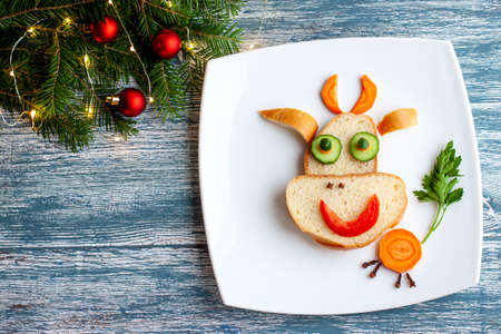 A perky bull from white bread, carrots, cucumbers, parsley as a Breakfast decoration for a child for Christmas and New year on a large white plate on a blue wooden background.の写真素材