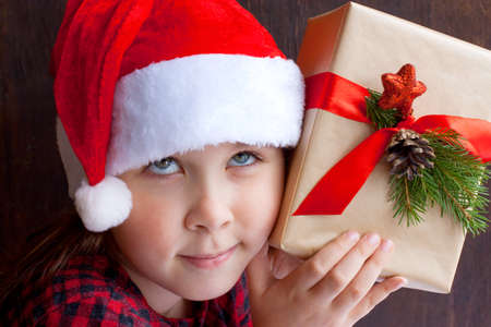 A close-up portrait of a girl in a red Santa hat and red shirt with her eyes up, holding a Christmas gift to her ear and listening to whats inside, against a dark wooden background Waiting a miracleの写真素材
