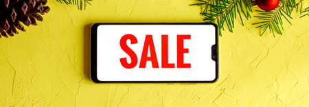 The inscription Sale in English on a white background of a black cell phone horizontally, lying on a yellow background and Christmas new year decor of fir branches and balls . Banner.の写真素材