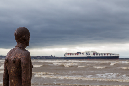 One of Antony Gormley's life-size statues of himself (which make up "Another Place" on the Sefton coastline) watches a container ship exiting the River Mersey.のeditorial素材