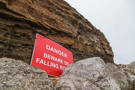 Danger beware of falling rocks sign pictured at the foot of the cliffs protecting the village of Staithes on the North Yorkshire coast seen in September 2020.の写真素材