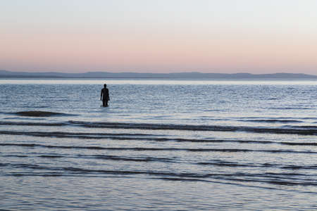 A lone Iron Man seen off the beach at Crosby at hight tide at dusk in April 2021.  He is part of Another Place, the art installation on Crosby beach near Liverpool made up of 100 Iron Men statues created by Antony Gormley.のeditorial素材