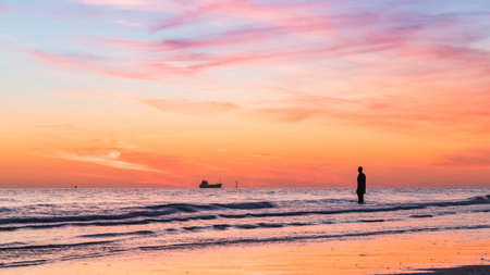 A lone Iron Man watches a large boat going out into the Irish Sea at sunset.  This is part of Another Place, the art installation on Crosby beach near Liverpool made up of 100 Iron Men statues created by Antony Gormley.  This was captured in April 2021.のeditorial素材