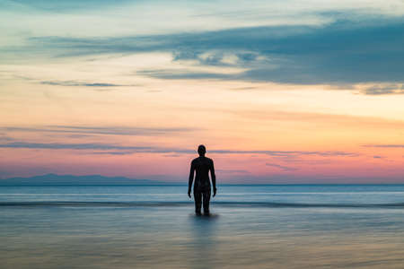 The tide rushes in surrounding an Iron Man at sunset in April 2021.  He is part of Another Place, the art installation near Liverpool made up of 100 Iron Men statues created by Antony Gormley.のeditorial素材