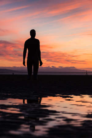 An Iron Man watches a spectacular sunset over the Irish Sea on Crosby beach near Liverpool in September 2021.  He is one of the 100 Iron Men statues created by Antony Gormley which occupy this stretch of the Sefton coastline.のeditorial素材