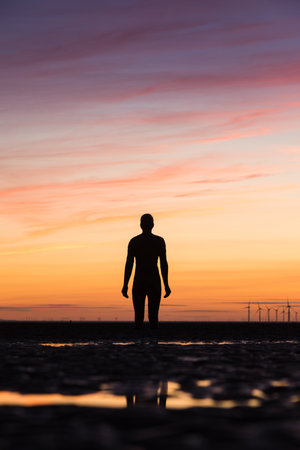 An Iron watches the sun setting behind the spinning windmills of Burbobank Windfarm off the Sefton Coast in Liverpool.  He is one of the 100 Iron Men statues created by Antony Gormley which occupy this stretch of Crosby beach near Liverpool.のeditorial素材