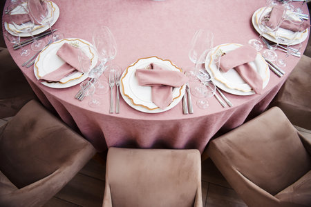 Beautiful table setting with white plates, golden forks, knives, empty glasses and pink napkins. Soft beige chairs adding elegance to interior. Festive celebration.の写真素材