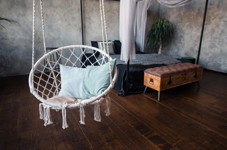 Comfy wickered chair hanging on ropes among bedroom with modern interior. Comfy bed, plants in pots and commode on background.の写真素材