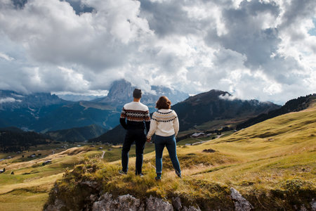 Back view of young man and woman holding hands and enjoying beautiful landscape of rocky mountains among cloudy sky. Active vacation at italian dolomites.の写真素材
