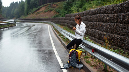 Young caucasian woman with backpack standing on wet road among mountains and using smartphone. Female hiker searching for route on cell phone.の写真素材