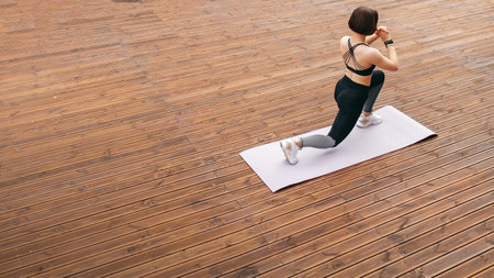 High angle view of sportswoman doing sit ups on fitness mat on wooden area. Back view of beautiful girl doing exercise in sportswear and sneakers. Concept of sport at outdoors. Daytime. Copy spaceの写真素材