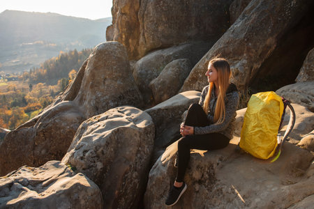 Outdoor recreation. Young girl in sportswear, in gray jacket and backpack sitting on stone mountain, looking away, in background autumn forest and mountains. Concept of tourism.の写真素材
