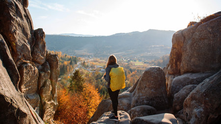 Outdoor recreation. Young girl in sports clothes, in gray jacket and backpack on her shoulders stands back, on stone mountain, in background small village in mountains. Autumn. Concept of tourism.の写真素材