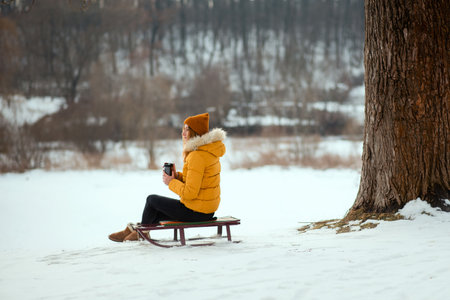 Young woman in yellow winter jacket sitting on a sleigh and drinking tea from the thermo cup in the winter park.の写真素材