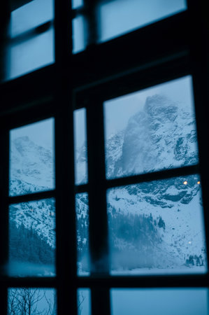 A serene winter landscape viewed through a window. Snow-covered mountains rise in the background, shrouded in mist. The scene is tranquil, with a cool blue hue dominating the atmosphere, suggesting a cold, peaceful day.の写真素材