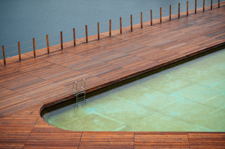 A wooden deck surrounds a partially filled swimming pool next to a body of water.