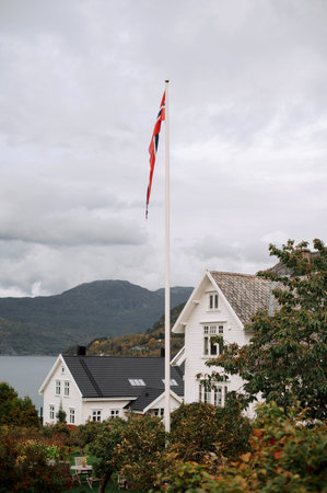 A scenic view of a white wooden house with a Norwegian flag on a pole, set against a backdrop of mountains and a lake under a cloudy sky.の写真素材
