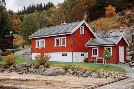 A charming red wooden house with white trim, surrounded by autumn foliage and rocky terrain. The house is situated near a sandy area, possibly a beach, with a backdrop of trees.の写真素材