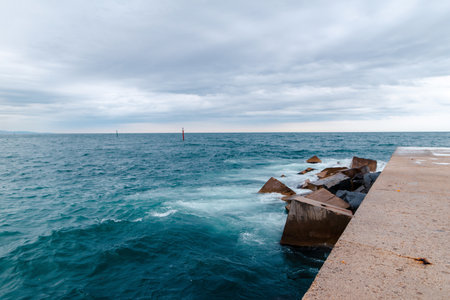 A concrete pier extends into a choppy sea under a cloudy sky, with waves crashing against large rocks.の写真素材