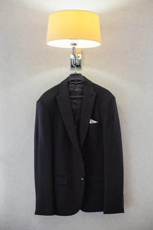 A black suit jacket hangs on a wall-mounted hanger beneath a lit lamp, with a white pocket square...