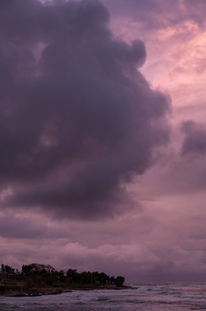 A dramatic seascape featuring dark clouds looming over a tranquil beach at sunset. The sky is...