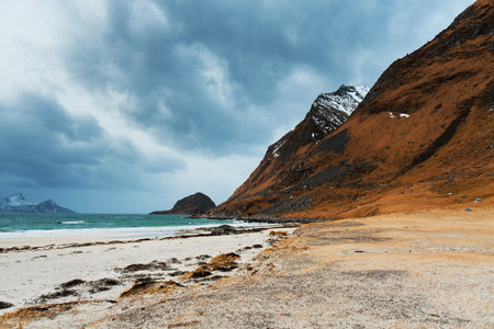 A scenic coastal landscape with a sandy beach, rocky mountains, and a cloudy sky.の写真素材