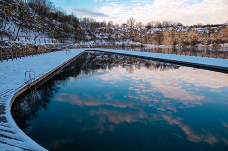 A serene winter scene of a natural pool surrounded by snow-covered cliffs and trees, reflecting the...