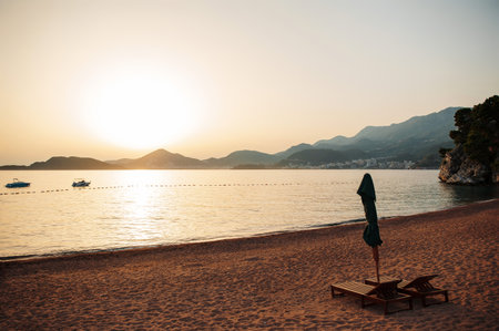 A serene beach scene at sunset with two empty lounge chairs and an umbrella on the sand. The calm sea reflects the golden hues of the setting sun, with distant mountains in the background.の写真素材