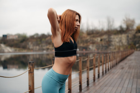 Fitness woman with red hair stretching outdoors on a wooden bridge near a lake. Girl wearing sporty black bra and leggings pantsの写真素材