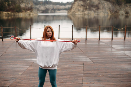 Red hair woman in a white hoodie and teal leggings exercises with a resistance band on a wooden deck by a lake. Girl training outdoorの写真素材