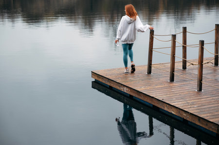 Red hair woman in sporty clothing stands on a wooden dock by a calm lake, holding a water bottle. The scene is serene and reflective, with the water mirroring the overcast sky.の写真素材