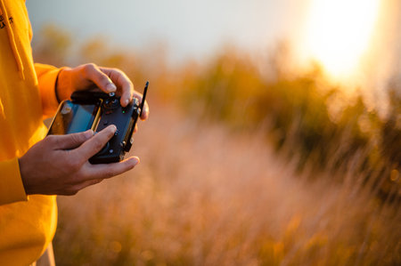 A person in a yellow hoodie operates a drone controller outdoors during sunset, with blurred grass and sunlight in the background.の写真素材