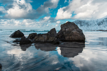 Rocky beach with reflections on wet sand, cloudy sky, snowy mountains.の写真素材