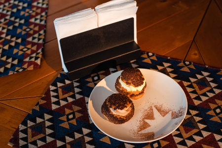 Two cream puffs with chocolate topping on a white plate with cocoa powder design, placed on a wooden...