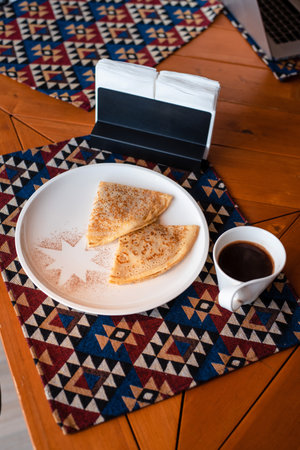 Two folded crepes on a white plate with cinnamon, next to a cup of coffee on a patterned tablecloth.