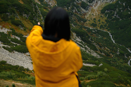 Black hair hiker woman in yellow jacket pointing at a mountainous landscape. Tatry, Polandの写真素材