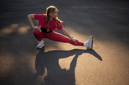 Cute blonde woman in pink sportswear stretching on asphalt road at sunny autumn day. Healthy and sporty conceptの写真素材