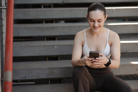 Young woman sitting on stairs, wearing a sports bra and leggings, smiling while using a smartphoneの写真素材