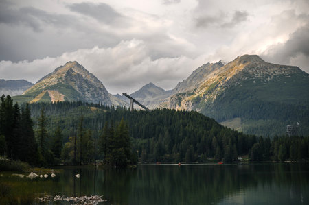 Scenic view of a tranquil lake surrounded by mountains and pine trees, with a ski jump structure in...