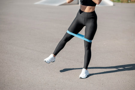 A woman performs a leg abduction exercise outdoors using a blue resistance band around her thighs to...