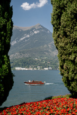 A classic wooden boat cruises across the serene waters of Lake Como, with lush green mountains and a charming town in the backgroundの写真素材