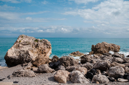 Large boulders and pebbles line the shore where the vibrant blue ocean meets the land under a cloudy...