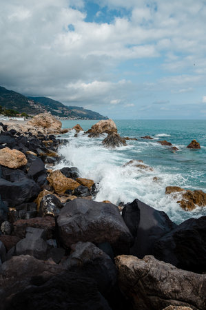The turquoise sea crashes against a rocky shore with large dark boulders and scattered lighter stones, under a cloudy sky with a distant coastal village nestled on a green hillsideの写真素材